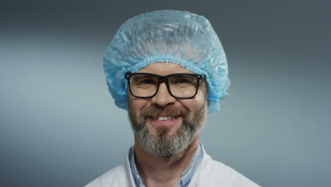 Portrait-of-the-Caucasian-handsome-grey-haired-man-physician-in-glasses,-blue-hat-and-with-a-beard-smiling-joyfully-to-the-camera.-Close-up.